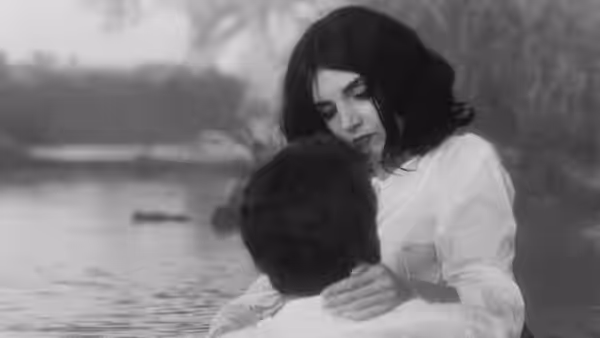 A woman rests her hands on a seated man's shoulders beside the water