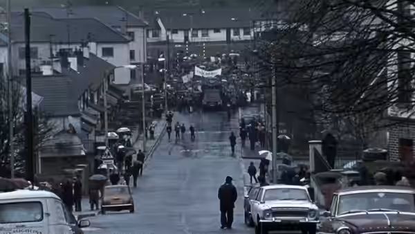 Marchers carrying banners walk down a residential street as people watch beneath umbrellas
