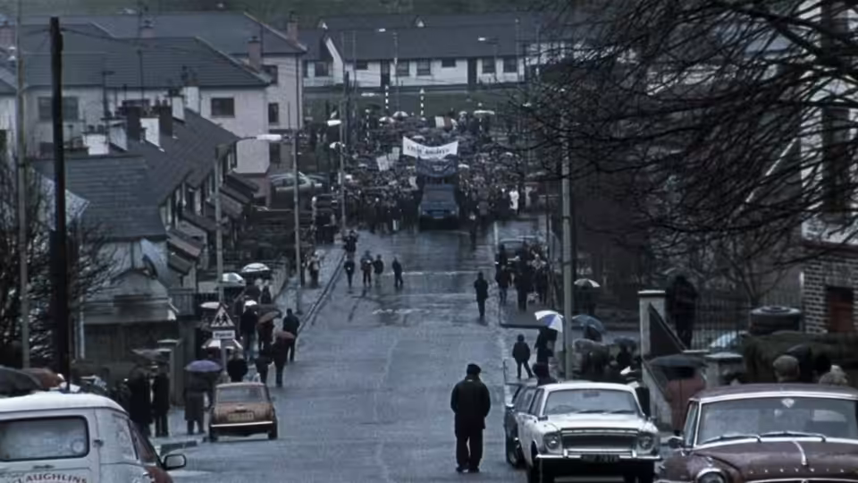 Marchers carrying banners walk down a residential street as people watch beneath umbrellas