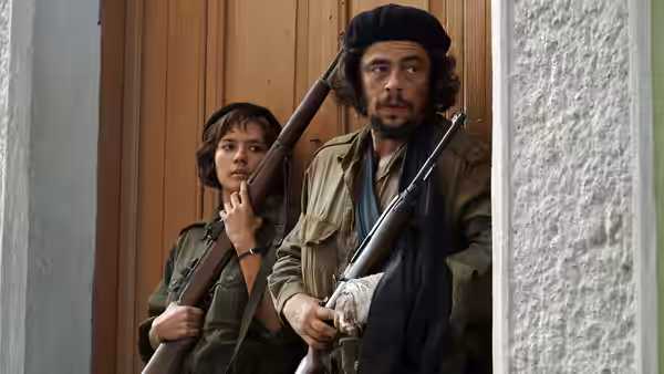 Ernesto Che Guevara (Benicio del Toro) holding a rifle in a doorway with a woman