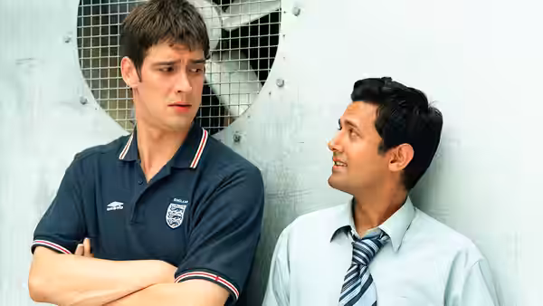 Two men standing against a wall one with arms crossed