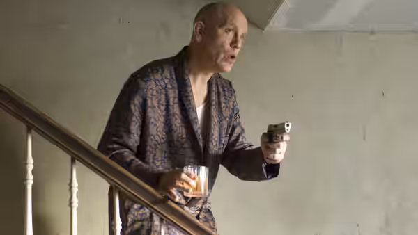 Osborne Cox (John Malkovich) stands on a staircase holding a drink and a handgun