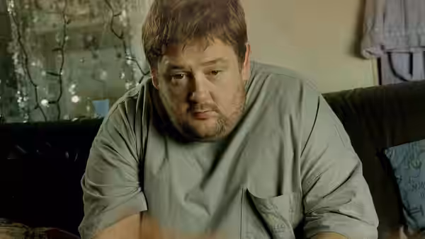 Moz (Johnny Vegas) seated on a couch looking down in a living room