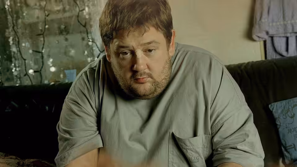 Moz (Johnny Vegas) seated on a couch looking down in a living room