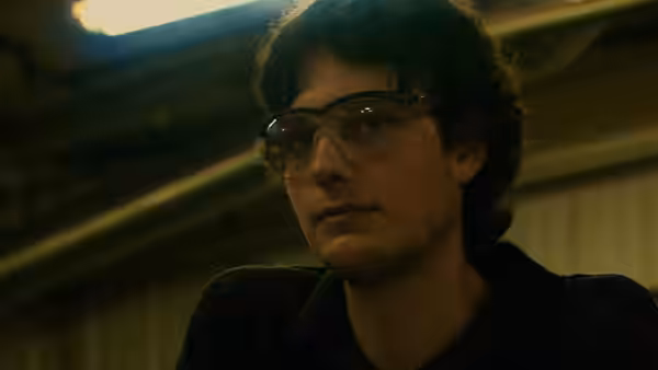 Young man in safety glasses and striped shirt looks to the side in a factory