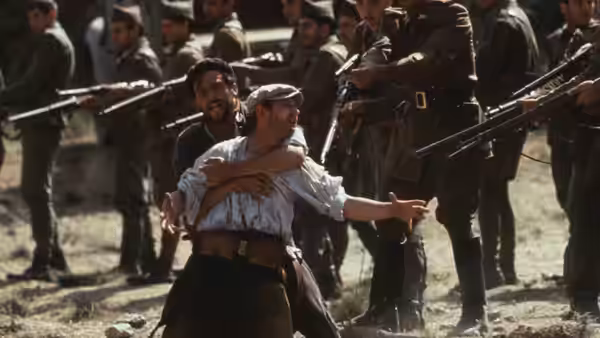 David Carr (Ian Hart) held by soldiers with rifles raised in a field