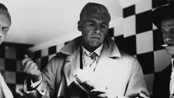 A man in a light trench coat stands between two men by a checkerboard wall