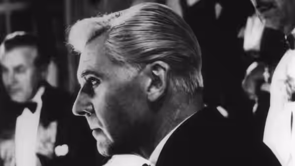 A silver-haired man in profile stands among formally dressed men