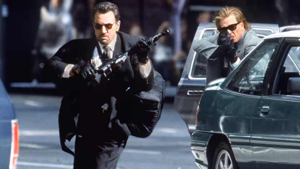 Lt. Vincent Hanna (Al Pacino) aiming a submachine gun beside a car