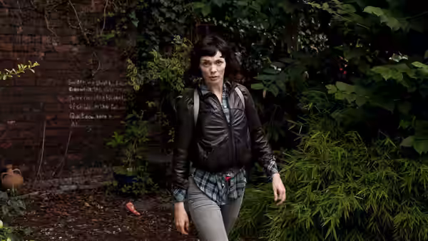 Jessica Hyde (Fiona O'Shaughnessy) walking past a brick wall in overgrown foliage