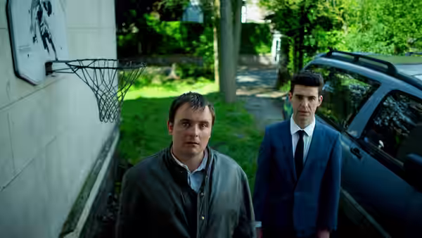 Two men standing beside a dark car outside a building with basketball hoop