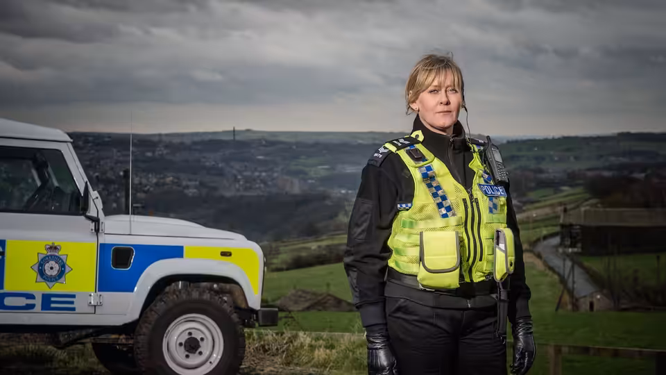 Catherine Cawood (Sarah Lancashire) stands beside a police vehicle in hilly countryside