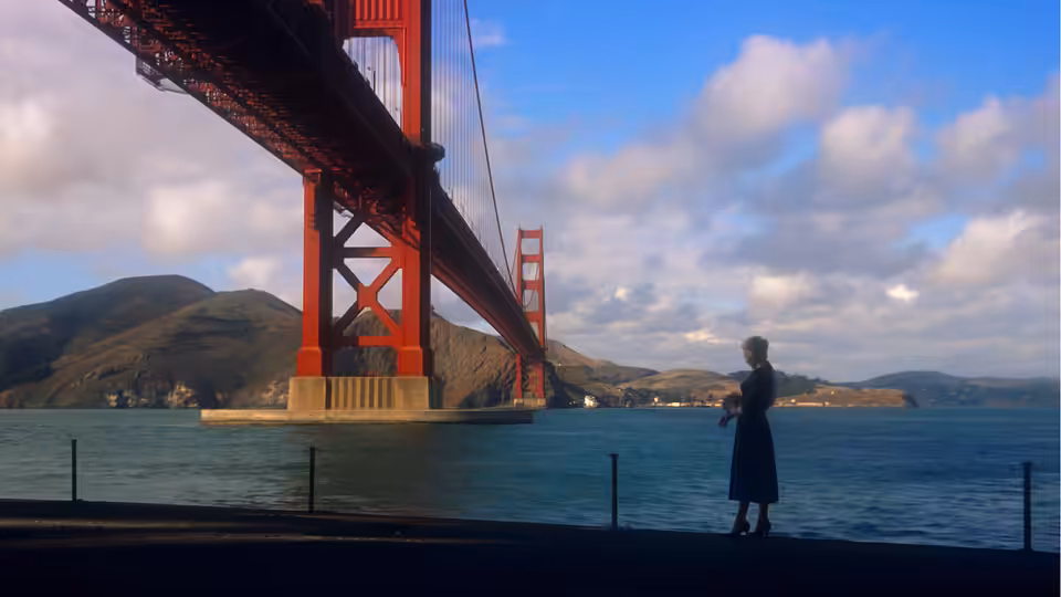a woman in a dark coat standing by the water under a red bridge