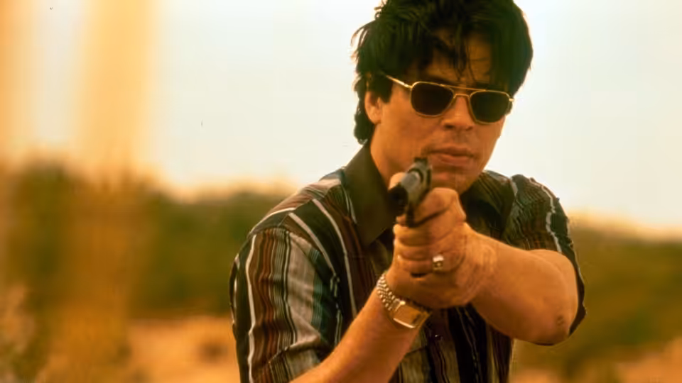 Javier Rodriguez (Benicio del Toro) aiming a pistol forward in daylight