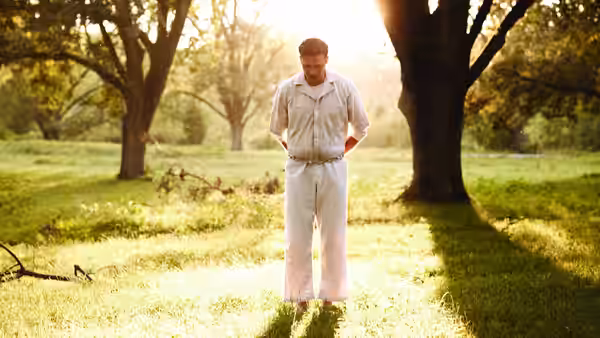 Man in light clothing stands in a sunlit meadow with trees behind him