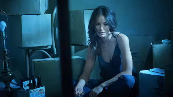 Woman in a blue dress sits among cardboard boxes holding a small object