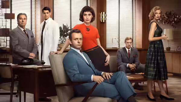 A man in a blue suit sits in an armchair surrounded by five colleagues in an office