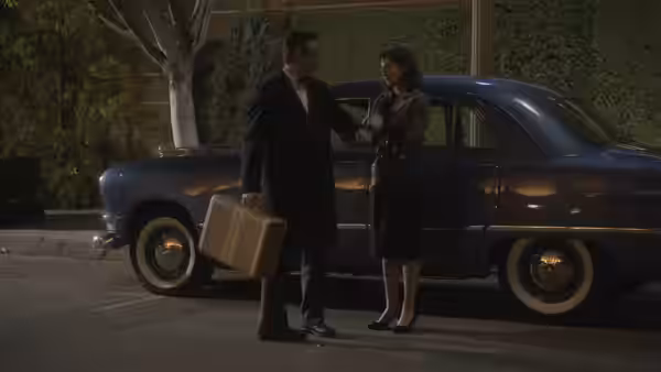 A man with a briefcase shakes hands with a woman beside a blue vintage car at night
