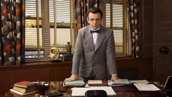 A man in a gray suit and bow tie stands behind a large cluttered wooden desk