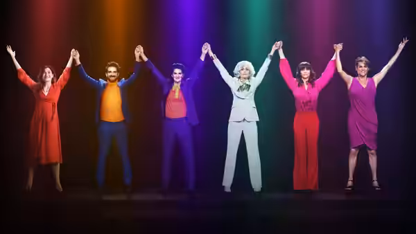 Six people in colorful outfits hold hands with arms raised on a rainbow stage