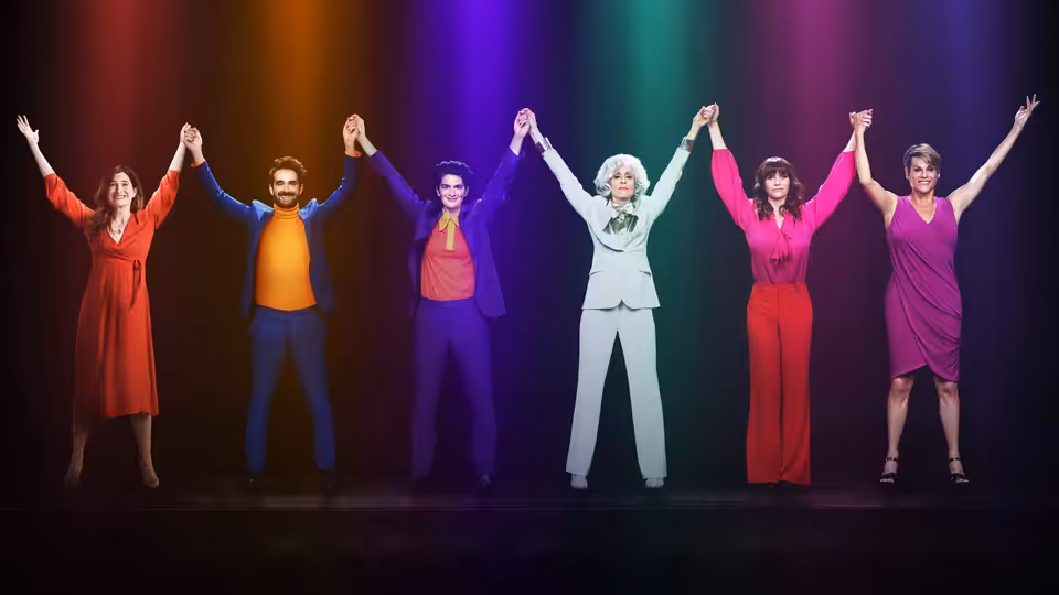Six people in colorful outfits hold hands with arms raised on a rainbow stage
