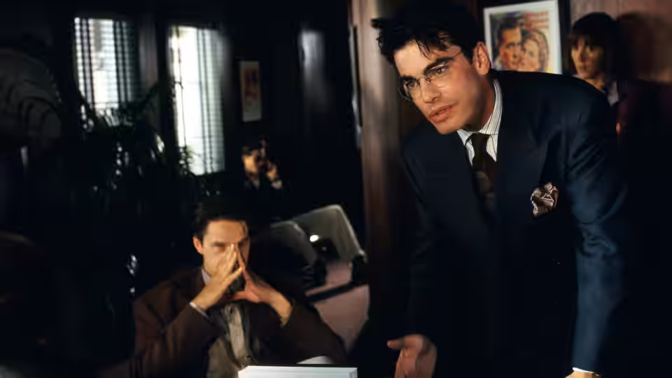 A suited man standing over another man seated in an office