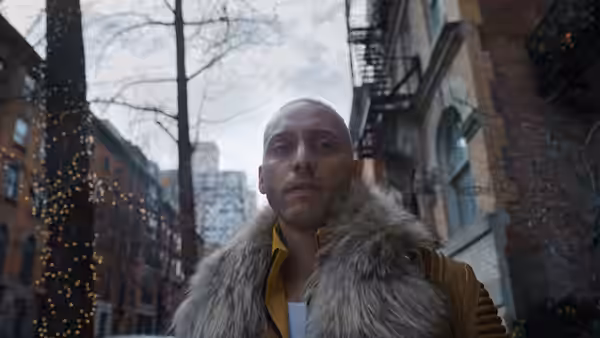 A bald man in a fur-collared jacket stands on a city street