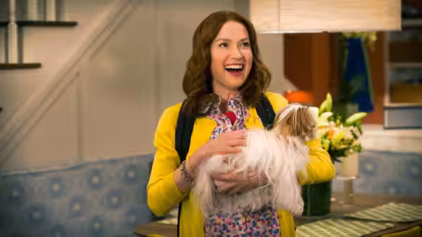 Kimmy Schmidt (Ellie Kemper) holding a small dog and smiling in a dining area