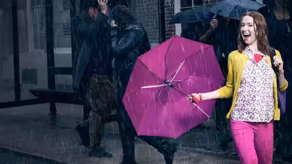 Kimmy Schmidt (Ellie Kemper) laughing with a pink umbrella in the rain on a city street