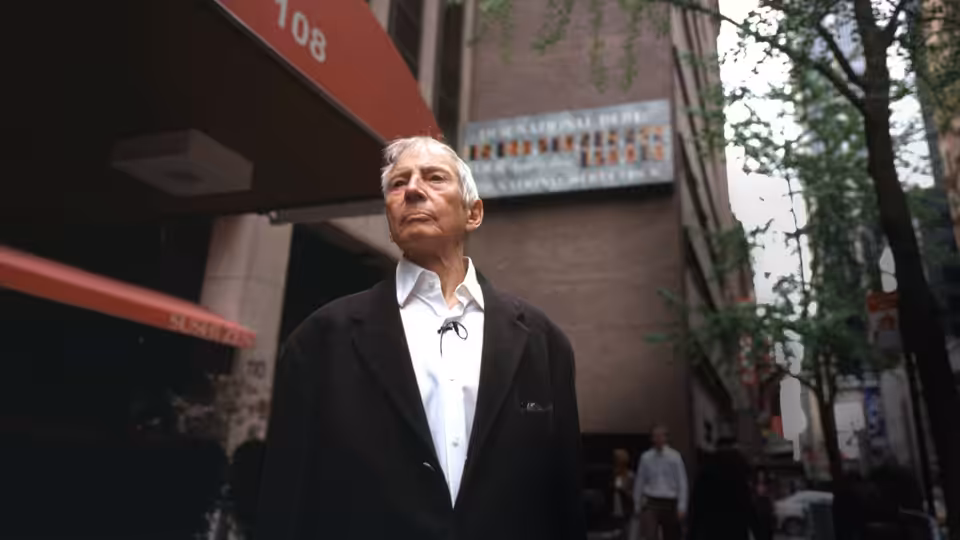 Robert Durst standing on a city sidewalk looking up at buildings