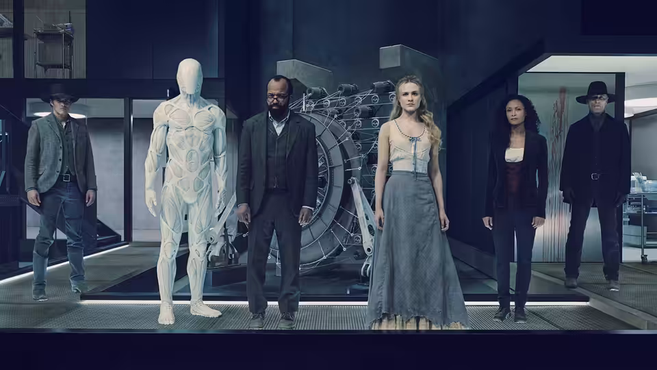 A blonde woman in a gray dress stands in a high-tech lab beside a white humanoid figure and suited men
