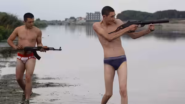 Two young men in underwear carry rifles by the water's edge