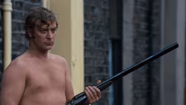 Shirtless Jack Carter (Michael Caine) holding a rifle outside a building