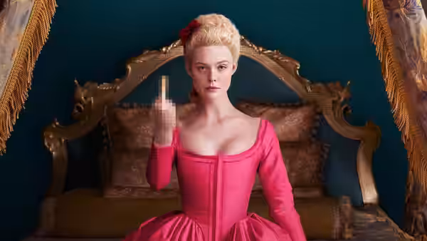 Catherine (Elle Fanning) sitting on a bed in a pink dress holding up one finger