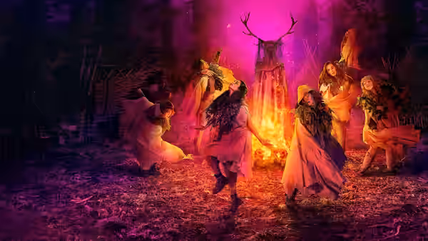 Women in flowing dresses dance in a circle around a fire with a deer skull