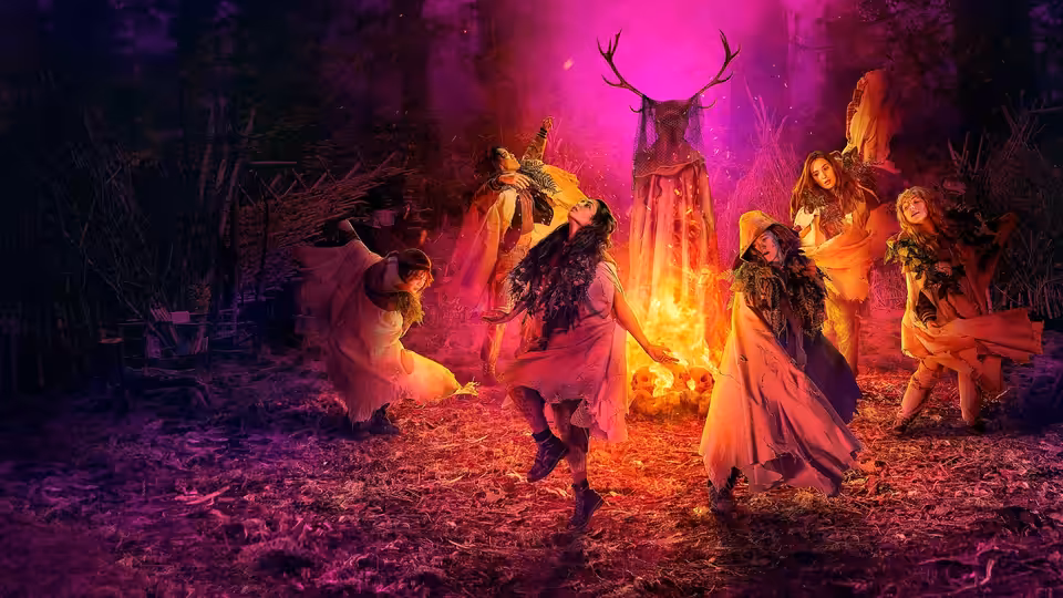 Women in flowing dresses dance in a circle around a fire with a deer skull