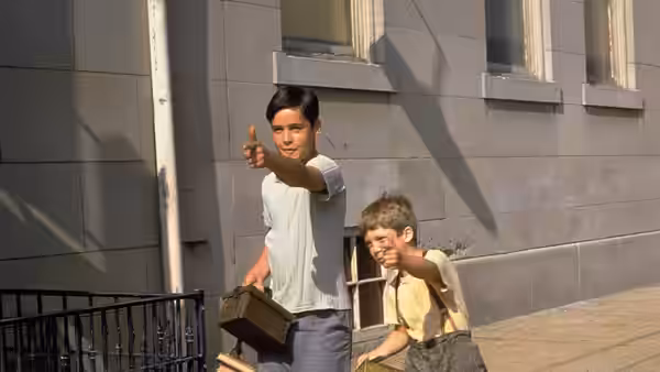 Two boys point ahead while holding suitcases outside a stone building