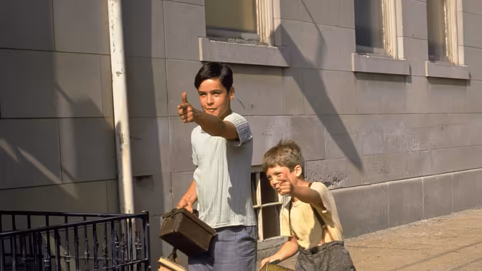 Two boys point ahead while holding suitcases outside a stone building