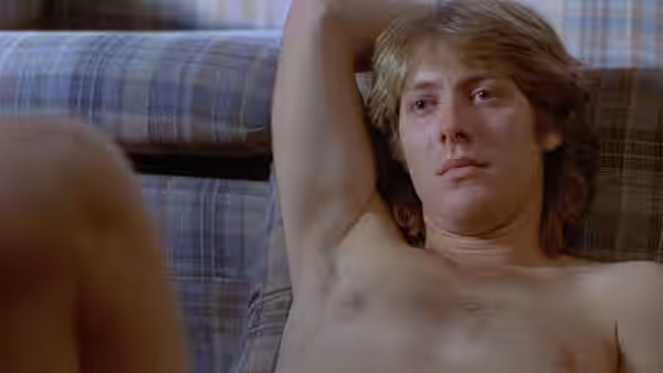 a shirtless blond man reclines on a plaid couch