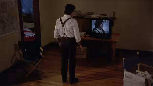 a man in suspenders watches a woman on a television screen