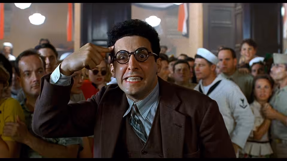 Barton Fink (John Turturro) gesturing with hand to head in crowded hall