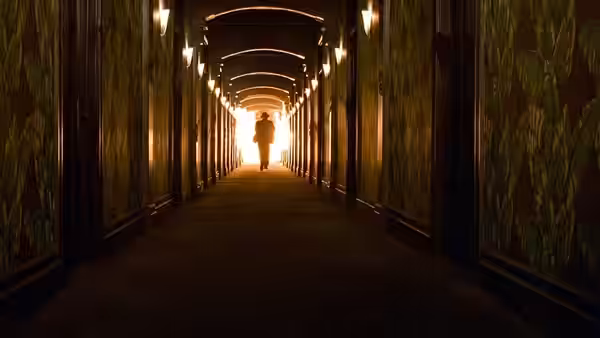 Silhouetted figure walks down long patterned hallway toward bright exit