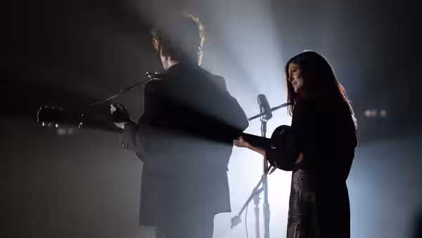 Joan Baez (Monica Barbaro) sings with a guitarist on a dark stage