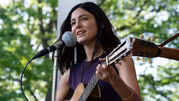 A woman sings into a microphone while playing acoustic guitar outdoors