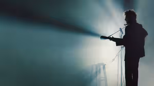 A guitarist performs on stage silhouetted against bright spotlight beams