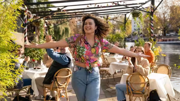 Jessie (Rose Matafeo) with arms outstretched at an outdoor cafe