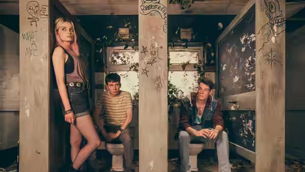 Maeve (Emma Mackey) standing as Otis (Asa Butterfield) and Adam sit on toilets