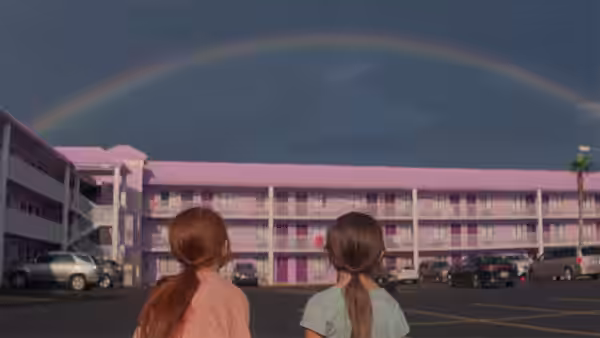 Two girls stand in a parking lot gazing at a rainbow over a pink motel