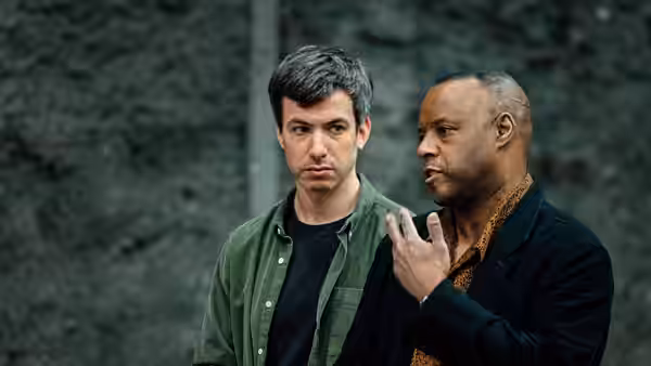 Nathan Fielder listens as another man speaks beside a stone wall