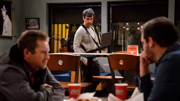 Nathan Fielder with laptop approaches two men seated at a table
